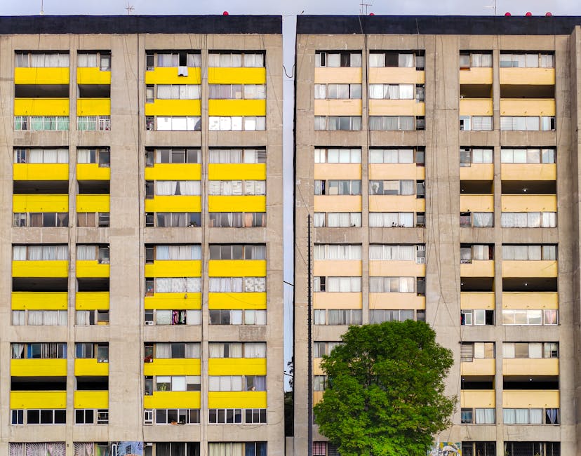 A photograph of a multi-storey residential building with a concrete facade, featuring numerous windows and balconies. The balconies have alternating yellow and beige window shutters, with some open or closed, and a few with visible laundry or curtains. The building is divided into two sections by a vertical gap, with the left side displaying more yellow window shutters. A large green tree is situated at the base of the building, partially obstructing the lower part of the structure. The scene is illuminated by daylight, showing a typical urban environment. This image relates to house removals and relocation services, as visualized by the setting and the context of moving logistics in high-rise residential buildings, emphasizing the challenges in access and how removals teams organize the transfer of furniture and boxes during home relocation.