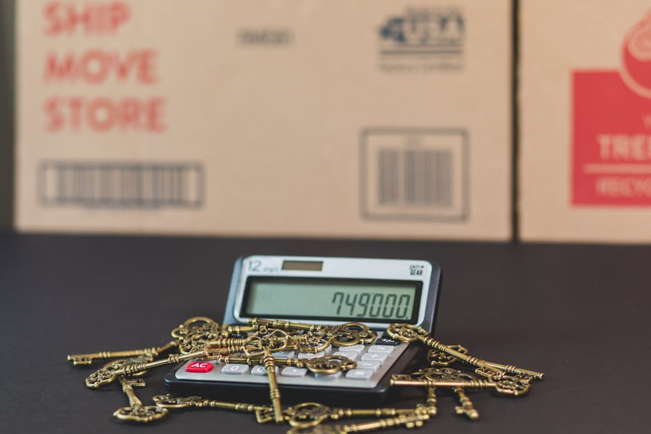 A close-up image displaying a calculator with a digital readout of 749,800 placed on a dark surface, surrounded by a pile of gold-colored antique keys of various sizes and shapes, some with ornate bows. Behind the calculator and keys, there are several large cardboard boxes stacked, with printed labels and barcodes indicating packaging for moving or storage. The boxes are positioned inside a room or warehouse environment, with natural or artificial lighting illuminating the scene. The image context suggests packing and logistics involved in home relocation, with a focus on the importance of organized packing materials and secure transport during house removals or furniture moving services, as provided by Removal Company Ilford.