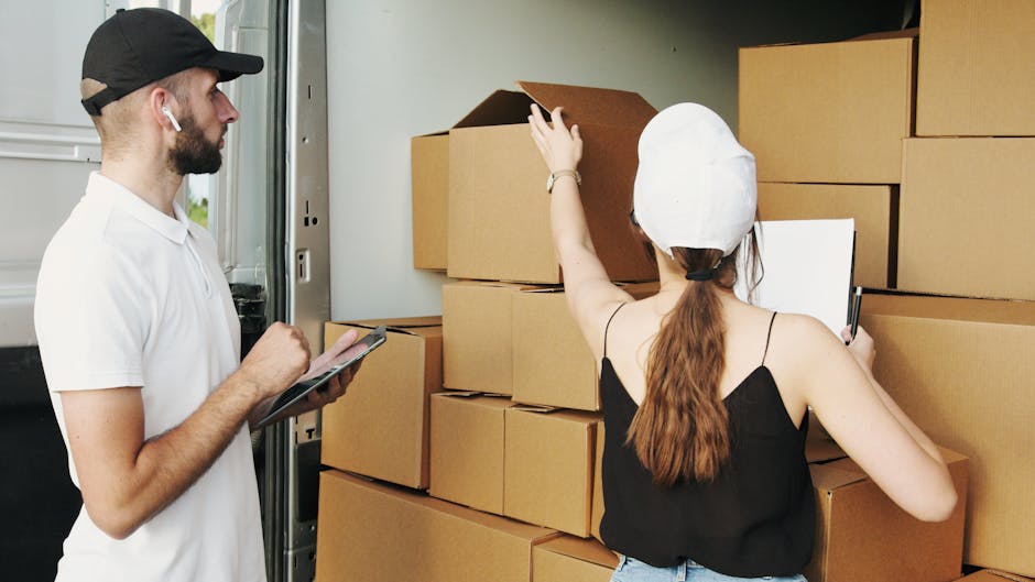 A male removal specialist wearing a white polo shirt, black cap, and wireless earbuds is standing next to a moving van, holding a clipboard and pen. A female assistant, dressed in a black sleeveless top and white protective cap, is inside the van and is reaching up to organize or check a stack of cardboard boxes. The boxes are various sizes, some open and some closed, and are packed with packing materials such as bubble wrap or paper. The van's interior is well-lit, showing the entrance with a metal threshold and the empty cargo space filled with neatly arranged boxes on a wooden surface. Outside, the surroundings suggest a residential area with a pavement and greenery. This scene captures the loading process during a home relocation, highlighting furniture transport and packing activities involved in professional removals services offered by Removal Company Ilford, as part of an organized moving or packing operation.