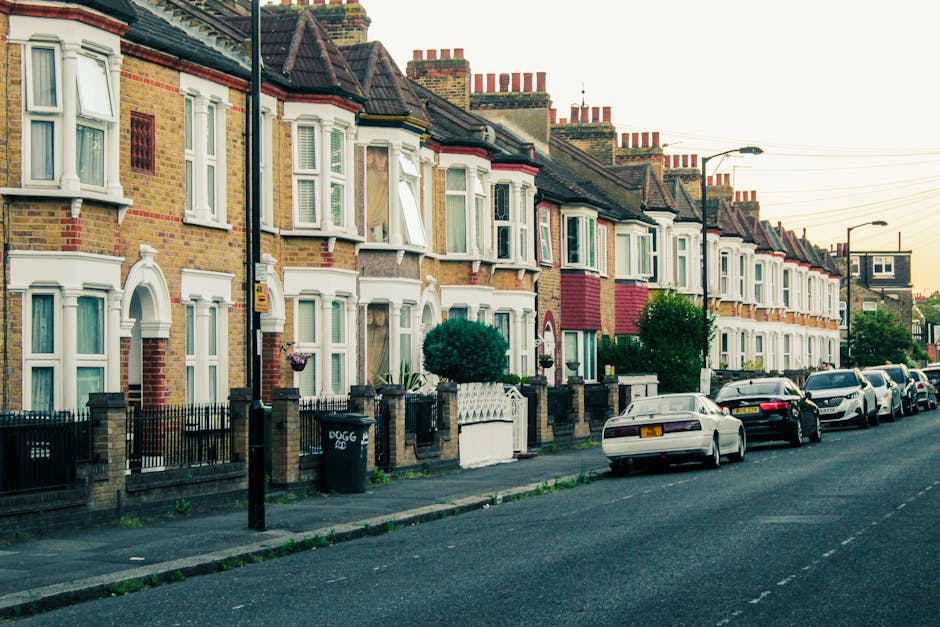 A row of traditional terraced houses on Ilford High Road during daylight, with some windows open and a mix of brickwork and painted facades. There are small front gardens bordered by low brick walls a