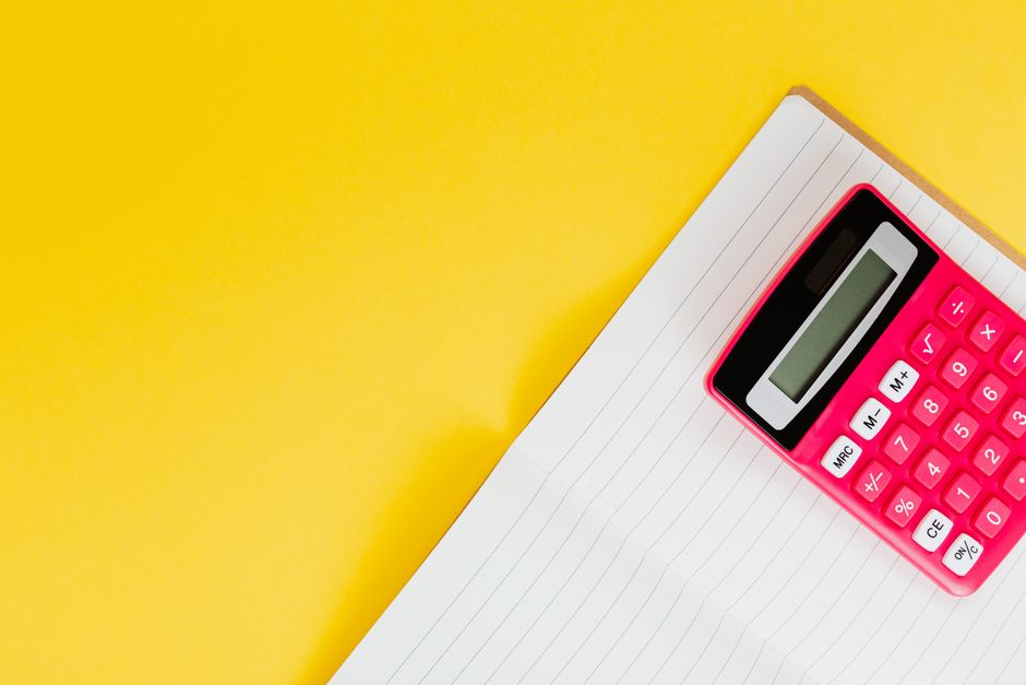 A bright pink calculator with a digital display and white buttons featuring mathematical symbols, placed on an open lined notepad with blank pages. The notepad is positioned on the corner of a desk or