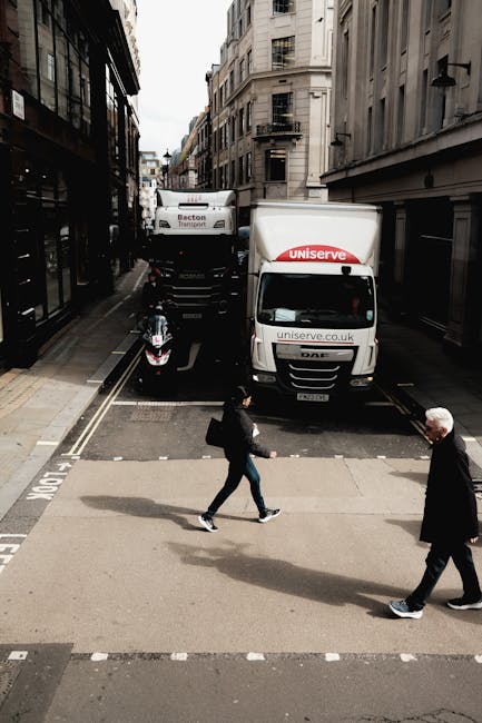 A view of a narrow city street captured from an elevated angle during daytime, with two small delivery trucks parked parallel to the curb near the entrance of a building. The trucks belong to Uniserve, a logistics company, and are positioned to facilitate home relocation or furniture transport as part of a moving process. In front of the trucks, there is a black scooter parked on the road, likely used for quick transport or delivery tasks. On the street, two pedestrians are crossing the road; one is a woman dressed in casual dark clothing carrying a bag, and the other is an older man with white hair wearing a dark jacket and trousers. The scene includes surrounding multi-storey buildings with varying architectural styles and black fire escape staircases, typical of urban residential areas. The street is lined with pavement, and the lighting suggests a partly cloudy day, providing a neutral and realistic view of a busy city environment involved in packing and moving logistics, with Removal Company Ilford providing the transportation services.