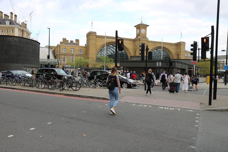 A busy scene outside Seven Kings station in ILFORD, featuring a large crowd of pedestrians crossing the street and walking on the pavement. Several people are carrying luggage or backpacks, indicating a mix of commuters and travelers. In the foreground, a woman in light blue jeans and a dark jacket is walking towards the station entrance. To the left, a row of parked bicycles is secured to a metal bike rack, with some bicycles partially covered by a black metal structure. The station itself is a prominent stone building with large arched windows and a clock tower displaying the time, with UK flags flying on top. Multiple traffic lights are visible, with red signals for vehicles and pedestrians. Nearby, a black van and other vehicles are either parked or waiting for the light to change, possibly in the process of loading or unloading items during a home relocation or furniture transport operation, indicating the involvement of ILFORD-based removal services such as those provided by Removal Company Ilford. The weather appears overcast with diffuse daylight illuminating the scene.