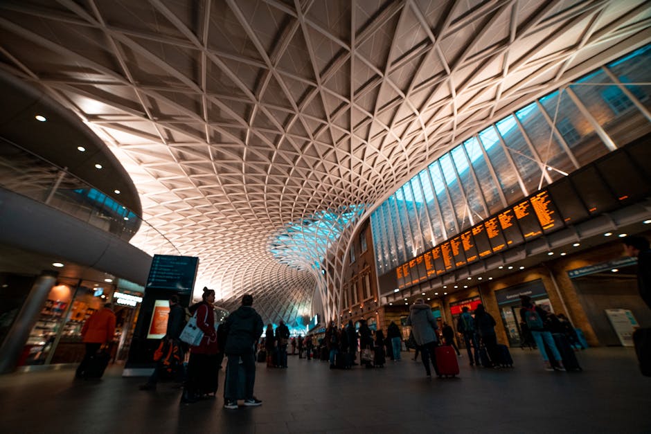 Inside Seven Kings station, the image captures a spacious, modern interior with a high, geometric ceiling made of interlocking wooden or metal panels arranged in a repeating diamond pattern, illuminated by overhead lighting. Large glass windows allow natural light to filter into the station, casting a subtle blue hue over parts of the space. Several commuters are visible, some standing with their luggage, others walking through the station. In the foreground, individuals are seen near a digital departure board displaying train schedules, and a few are holding or pulling wheeled suitcases and travel bags. The scene includes minimal furnishings such as seating and informational signs, with the background showing a corridor leading to platform areas. The environment is clean and well-organized, typical of a busy transport hub. As part of a house removal or furniture transport process, a removal company like Removal Company Ilford may be involved in assisting with luggage or equipment transfer within such transit environments, ensuring efficient packing and loading for home relocation or moving services.