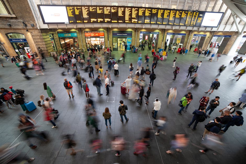 A busy scene outside Seven Kings station in ILFORD, featuring a large crowd of pedestrians crossing the street and walking on the pavement. Several people are carrying luggage or backpacks, indicating a mix of commuters and travelers. In the foreground, a woman in light blue jeans and a dark jacket is walking towards the station entrance. To the left, a row of parked bicycles is secured to a metal bike rack, with some bicycles partially covered by a black metal structure. The station itself is a prominent stone building with large arched windows and a clock tower displaying the time, with UK flags flying on top. Multiple traffic lights are visible, with red signals for vehicles and pedestrians. Nearby, a black van and other vehicles are either parked or waiting for the light to change, possibly in the process of loading or unloading items during a home relocation or furniture transport operation, indicating the involvement of ILFORD-based removal services such as those provided by Removal Company Ilford. The weather appears overcast with diffuse daylight illuminating the scene.