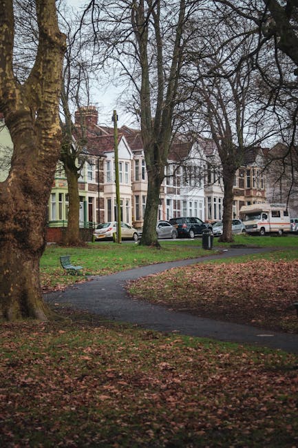 A paved pathway winding through a park area with leafless trees and patches of grass, situated in front of a row of multi-story Victorian-style houses with bay windows and brick façades. Several parked cars, including sedans and a van, are visible along the street near the houses. The scene is outdoors in natural daylight, with overcast skies creating diffused lighting. This setting illustrates a typical residential neighbourhood environment suitable for house removals and furniture transport, aligning with the context of home relocation services provided by Removal Company Ilford as described in the Valentines Park house removals tips article.