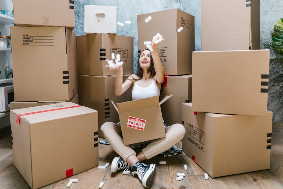 A young woman with long brown hair, sitting on a wooden floor surrounded by numerous cardboard removal boxes in a room. She is wearing a white sleeveless top, beige pants, and black and white sneakers. The woman is smiling and actively engaged in packing or unpacking, as she throws packing foam peanuts into the air from an open box in her lap. The room features a textured blue wall in the background, and a small plant is visible on the right side. Several boxes are stacked against the wall, with some sealed with red tape, indicating they are ready for moving or home relocation. Equipment such as a trolley or packing materials are not visible, but the scene suggests a busy packing process typical of furniture transport and house removals. The image illustrates the detailed packing stage of a professional moving service, consistent with home relocation activities handled by [COMPANY_NAME], supporting efficient and organized moving logistics during busy weekends, as referenced in the page titled 'Valentines Park house removals tips for busy weekends, ILFORD' on the removal company website.